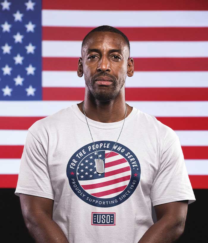 Image of service member wearing 2024 USO t-shirt standing in front of the American flag.