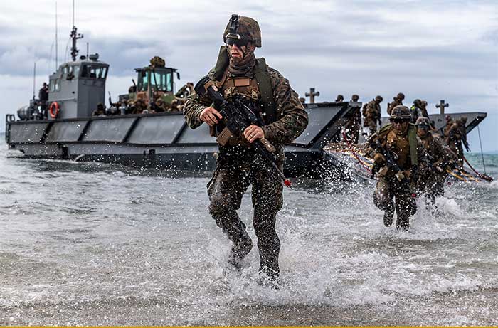 Marines run through the water with ship behind them.