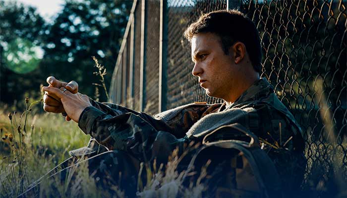 Service member stares at wedding band while sitting alone against a fence.