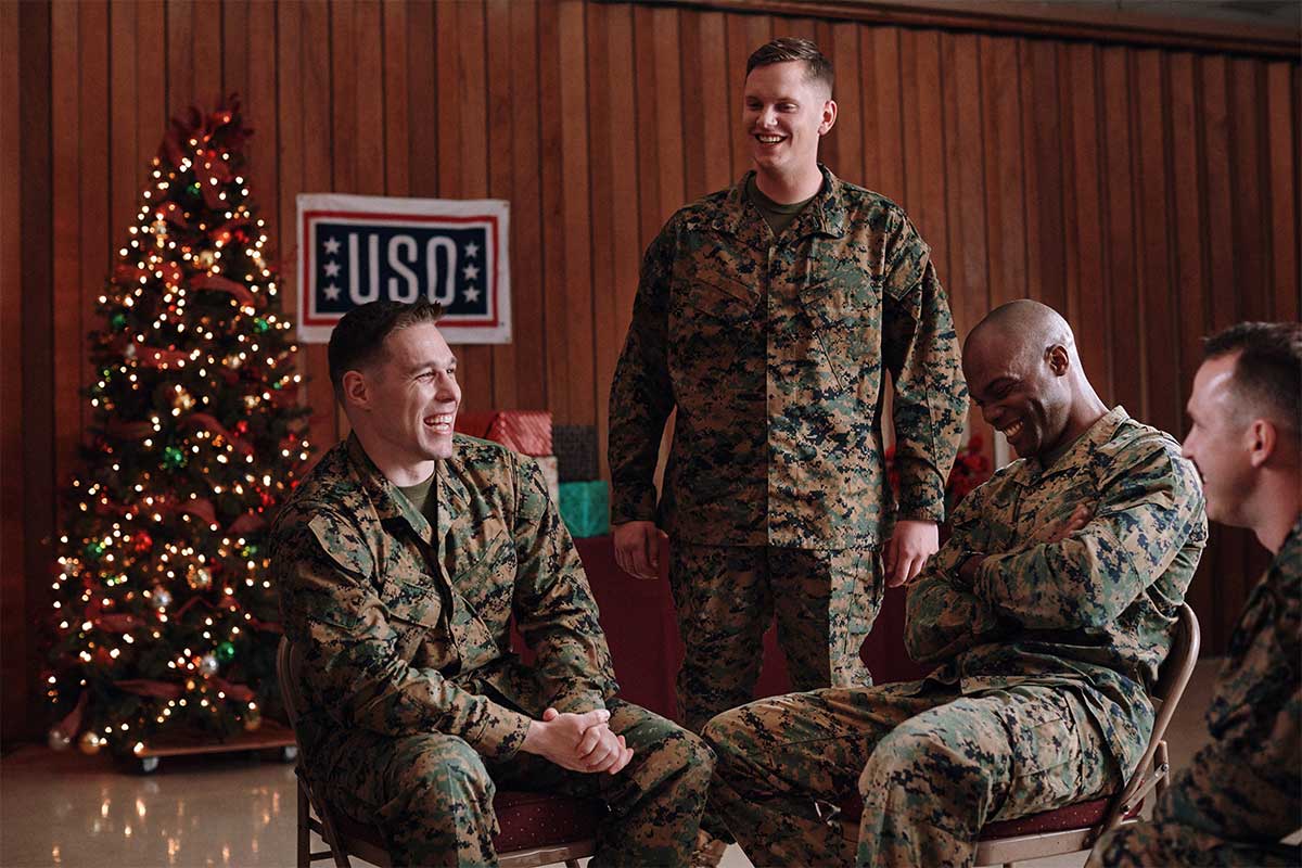 Four service members in uniform inside a USO Center in front of a Christmas tree and wrapped gifts