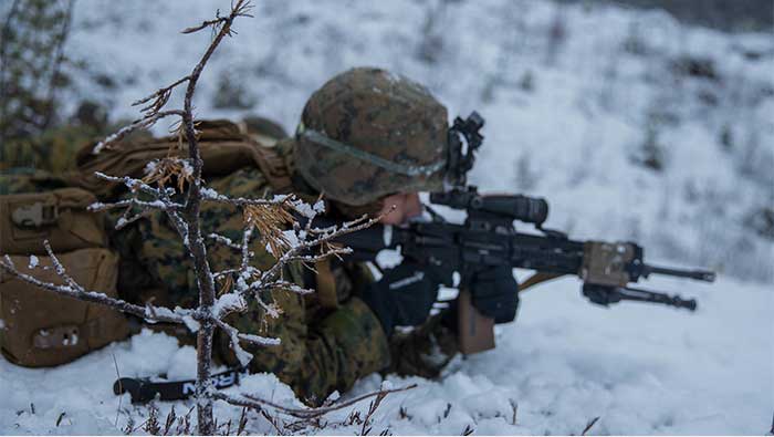 Service member aims gun in snowy terrain.